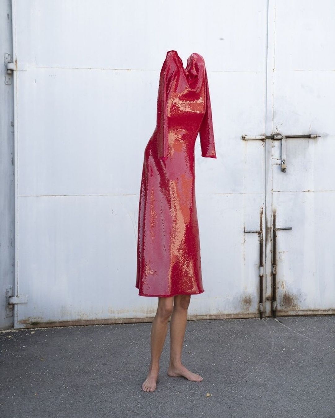 Red sequin dress on a mannequin with exposed legs, against a metal door and asphalt. Fashion editorial photography. Visual merchandising.