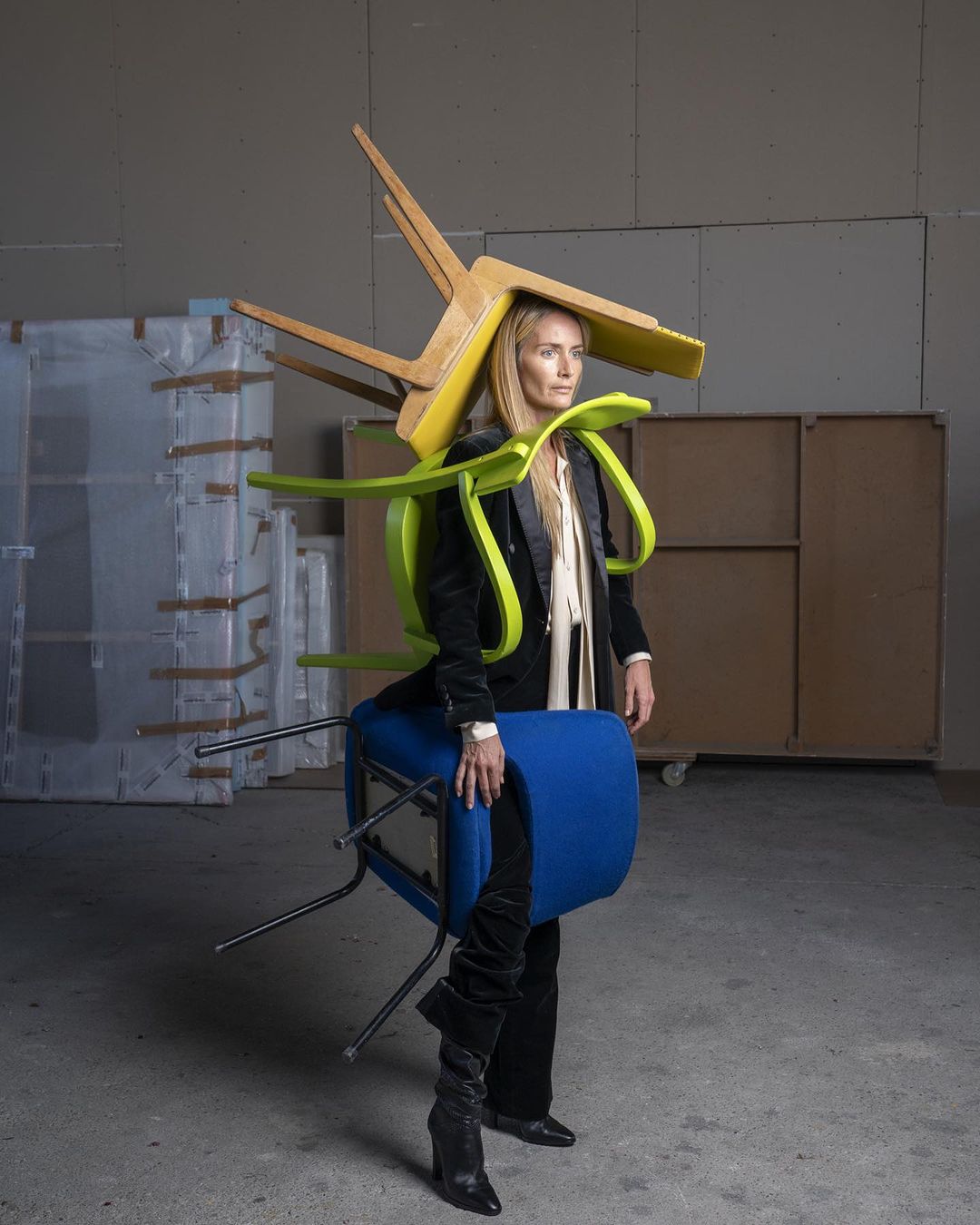 Fashion-forward portrait of a stylish woman wearing stacked colorful chairs as avant-garde couture in an industrial studio setting.