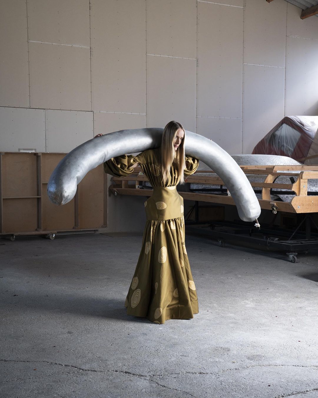 Woman in olive green dress carrying large, silver, curved pipe in an industrial-style room with concrete floor and light walls.