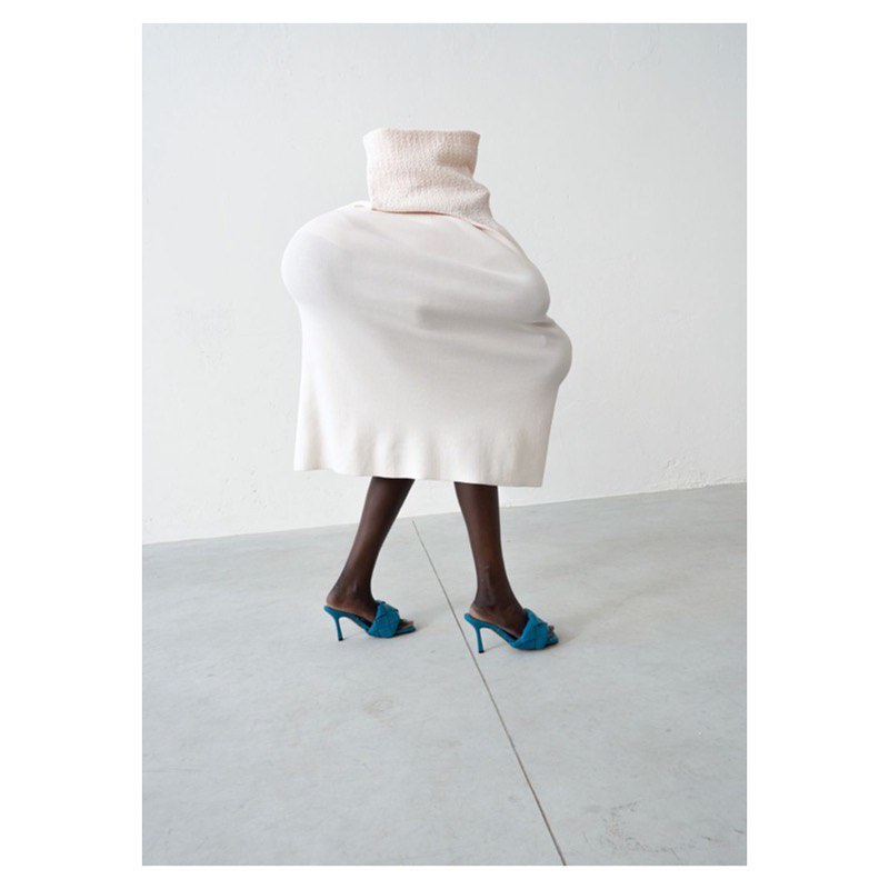 Fashion shot of a person wearing a large turtleneck sweater and a white skirt with blue heeled sandals in a white interior space.