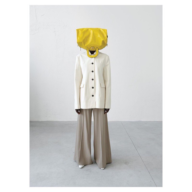 Full-length shot of a person wearing a white blazer and flared trousers, with a bright yellow bag covering their head in an indoor setting.