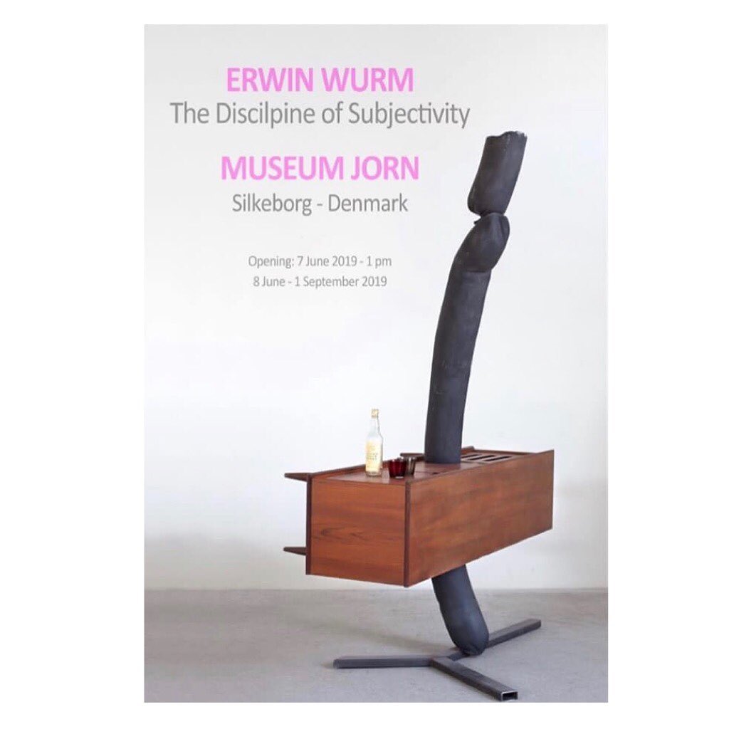 Abstract contemporary art piece featuring a table with a bottle and glass alongside a structural element in a bright art gallery setting.
