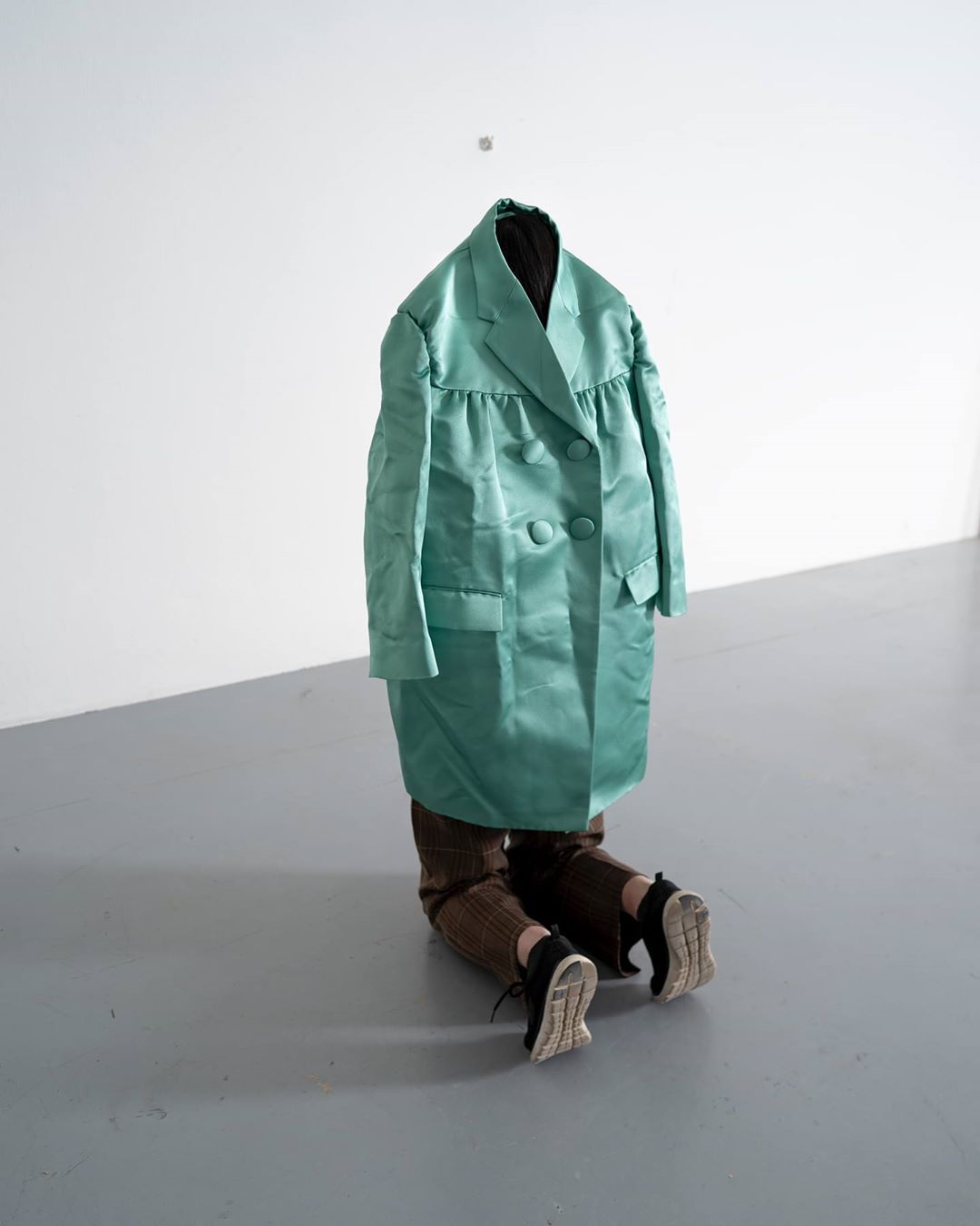 An adult kneeling with a teal, double-breasted coat draped over their upper body against a white wall. Styled for a fashion shoot.
