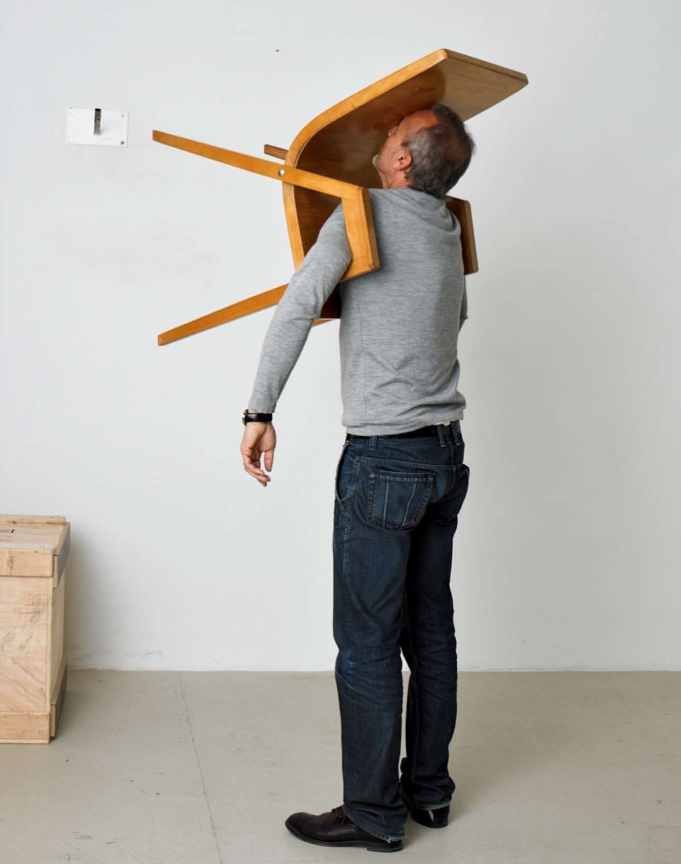 Man indoors with a wooden chair over his head, wearing a grey sweater and blue jeans, standing in front of a white wall.