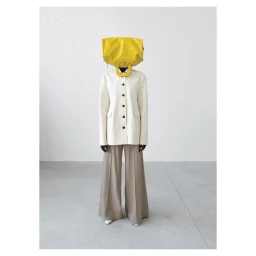 Model wearing a bright yellow handbag over their head, white buttoned jacket, and wide-leg trousers in a minimalist studio.