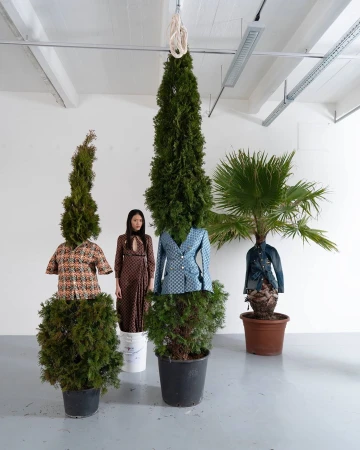 Three potted evergreen trees wearing patterned shirts and jackets alongside a standing woman in a patterned dress in a white studio