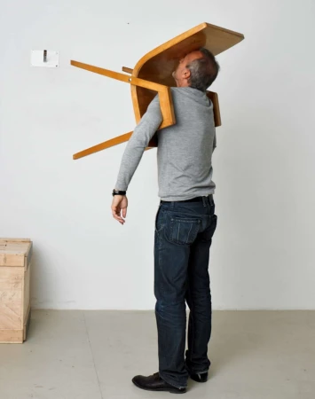 Man wearing grey long-sleeve shirt and dark jeans balancing a wooden chair on his torso against a white studio wall