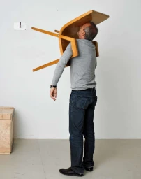 Man wearing grey long-sleeve shirt and dark jeans balancing a wooden chair on his torso against a white studio wall