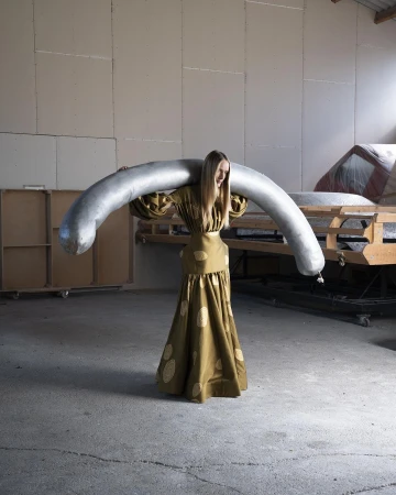 Woman in an olive dress holding a large silver sausage-shaped object across her shoulders in a warehouse