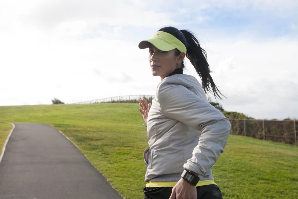 Athletic woman in a gray jacket and visor running on a path, looking over her shoulder in an outdoor setting with grassy hills.