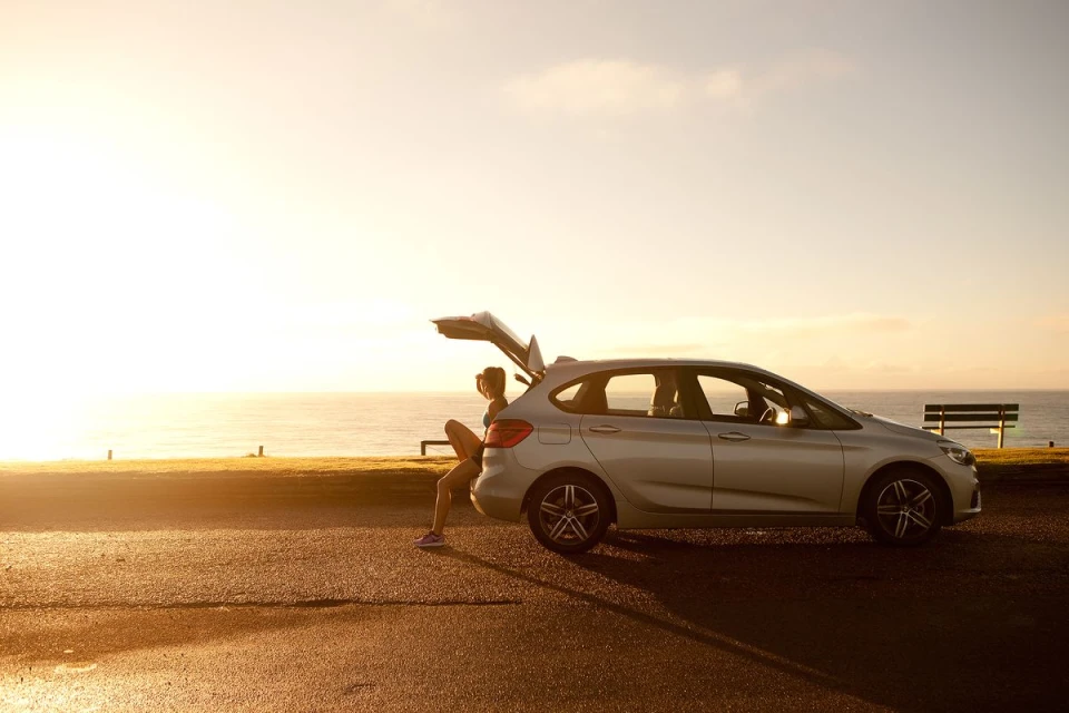 Silver hatchback parked along coast at sunset. A woman sits at the open rear hatch, enjoying ocean view. A bench is nearby.