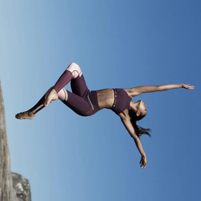 Athletic woman in fitness attire suspended against a blue sky with a rock edge visible, promoting an active, healthy lifestyle.