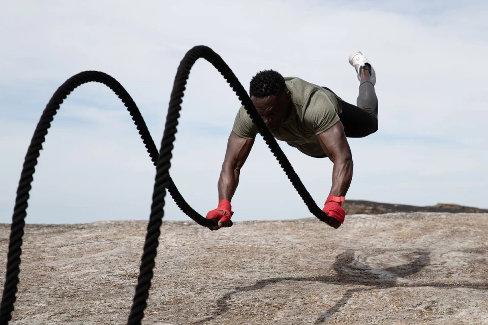 Athletic, muscular man wearing red gloves does pushups while suspended over rocky ground using thick, looped black battle ropes outdoors.