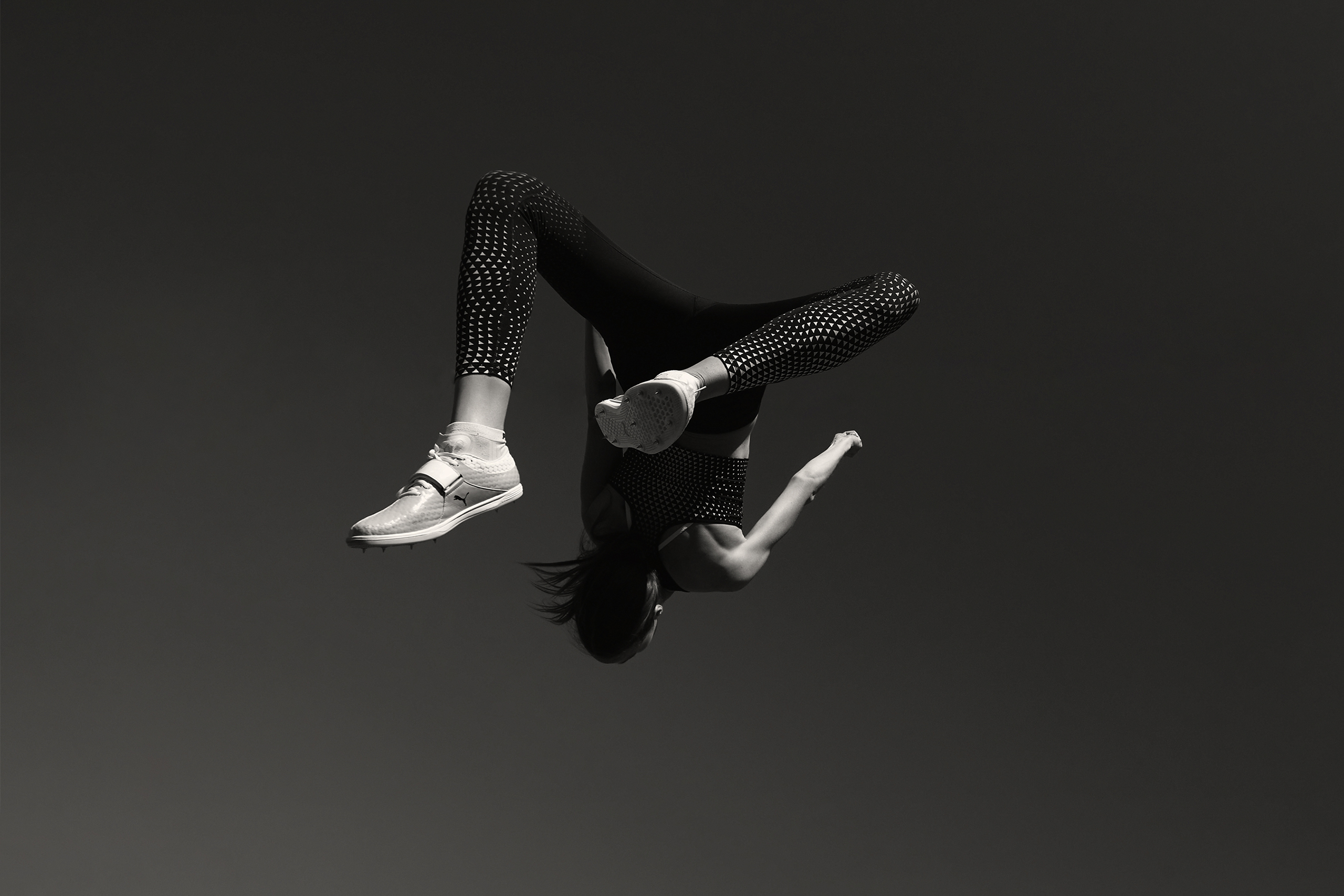 A woman in athletic wear mid-air performing a backflip in front of a dark gray background. She is wearing athletic shoes.
