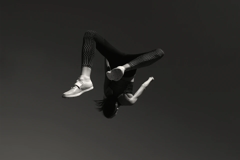 A woman in athletic wear mid-air performing a backflip in front of a dark gray background. She is wearing athletic shoes.