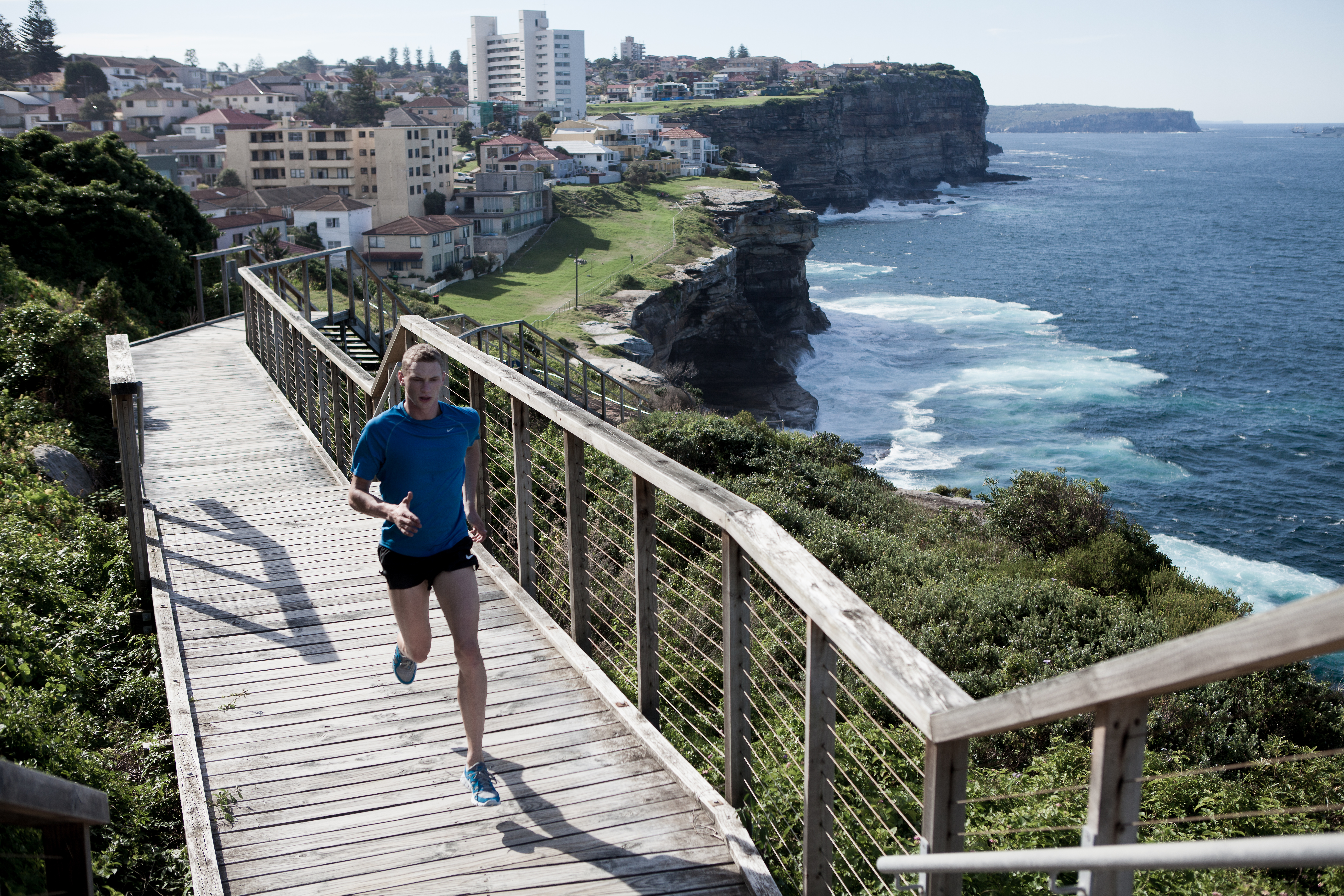 Male runner on wooden boardwalk with ocean and coastal cityscape backdrop, captured in a medium shot with clear lighting.