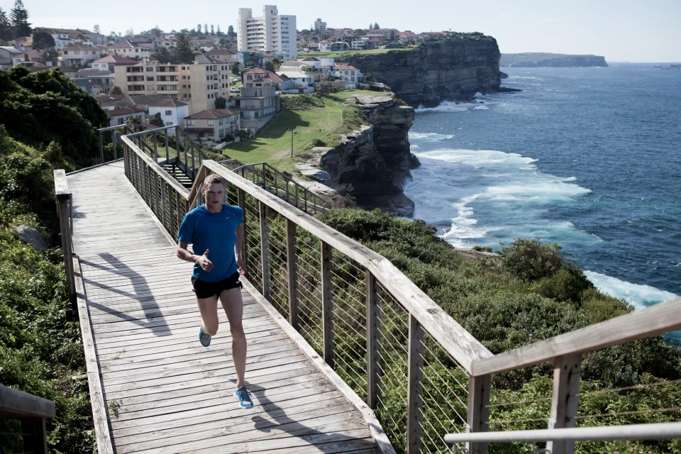 Male runner on wooden boardwalk with ocean and coastal cityscape backdrop, captured in a medium shot with clear lighting.