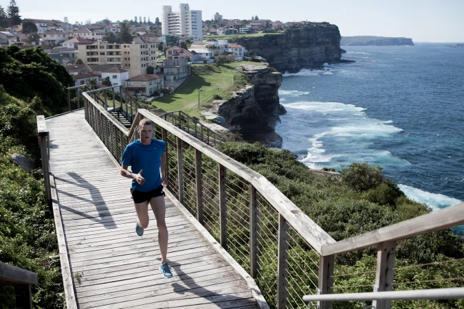 Male runner on wooden boardwalk with ocean and coastal cityscape backdrop, captured in a medium shot with clear lighting.