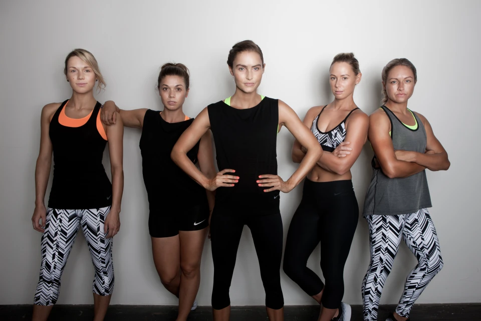 Five women pose in athletic wear in a studio setting, standing against a plain backdrop. They wear workout clothes. Fitness, healthy lifestyle.