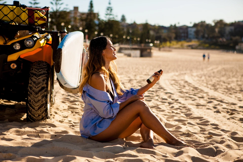 Woman sitting on sandy beach near ATV spraying sunscreen, wearing an unbuttoned blue shirt, with the ocean and city buildings visible.