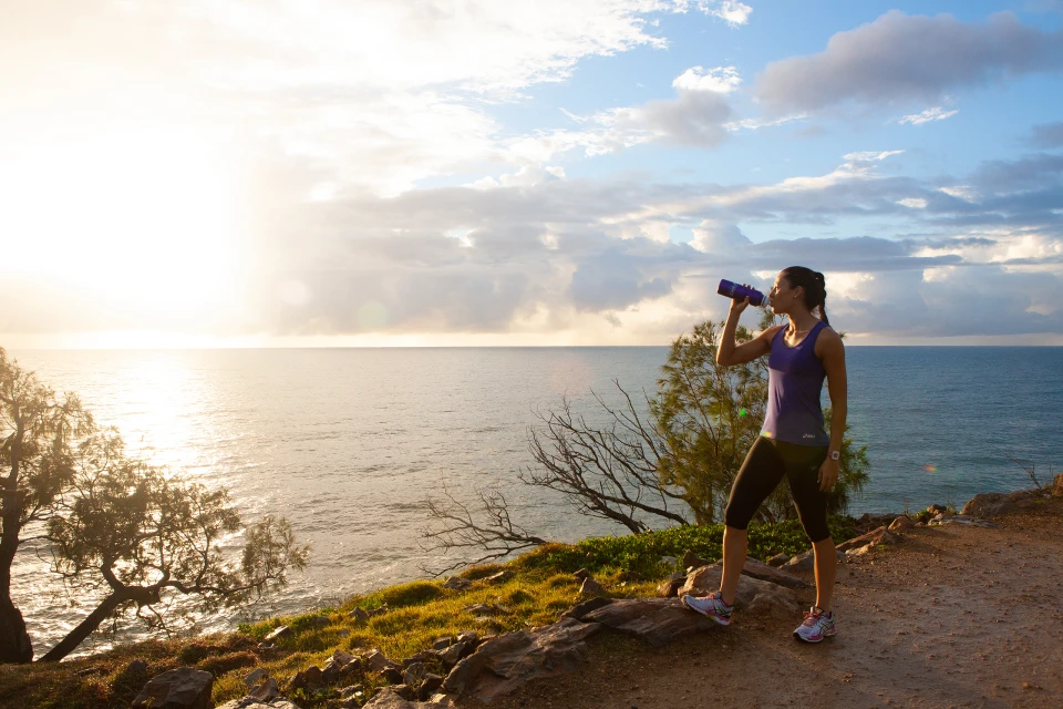 Woman drinks water bottle during an outdoor workout. Fitness shot on a hill overlooking the ocean, with trees and a cloudy sky.