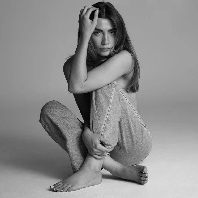 Black and white portrait of a young woman sitting on the floor in jeans, posing with hands on her head and legs, in a photo studio.