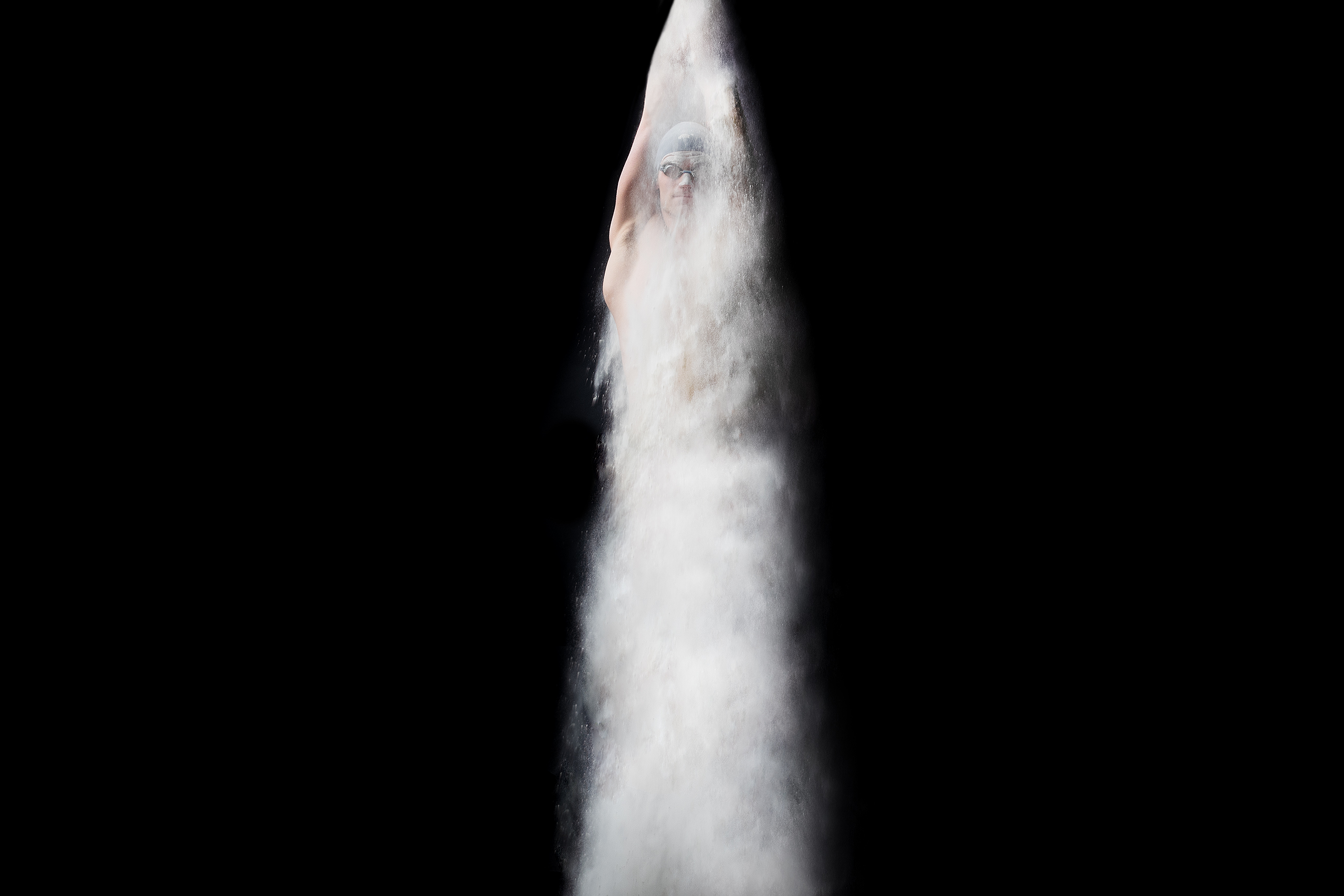 Adult swimmer wearing goggles emerging from a cloud of white powder in a studio against a black background, shot in portrait orientation.