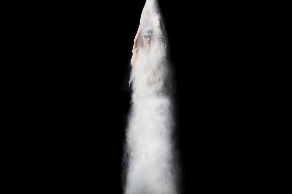 Adult swimmer wearing goggles emerging from a cloud of white powder in a studio against a black background, shot in portrait orientation.