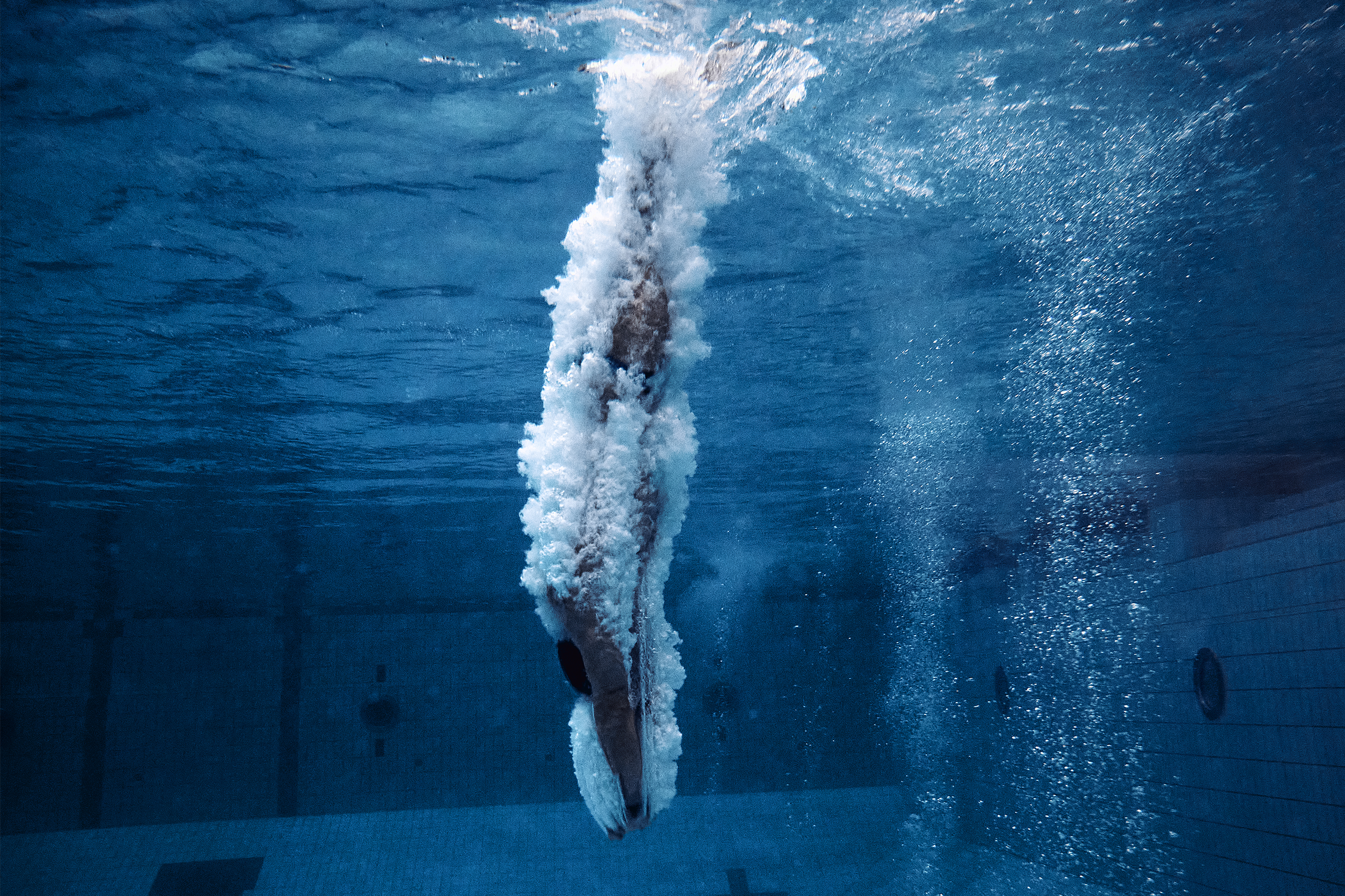 Underwater shot of a swimmer diving into a pool, surrounded by bubbles and blue water, capturing the moment of submersion.