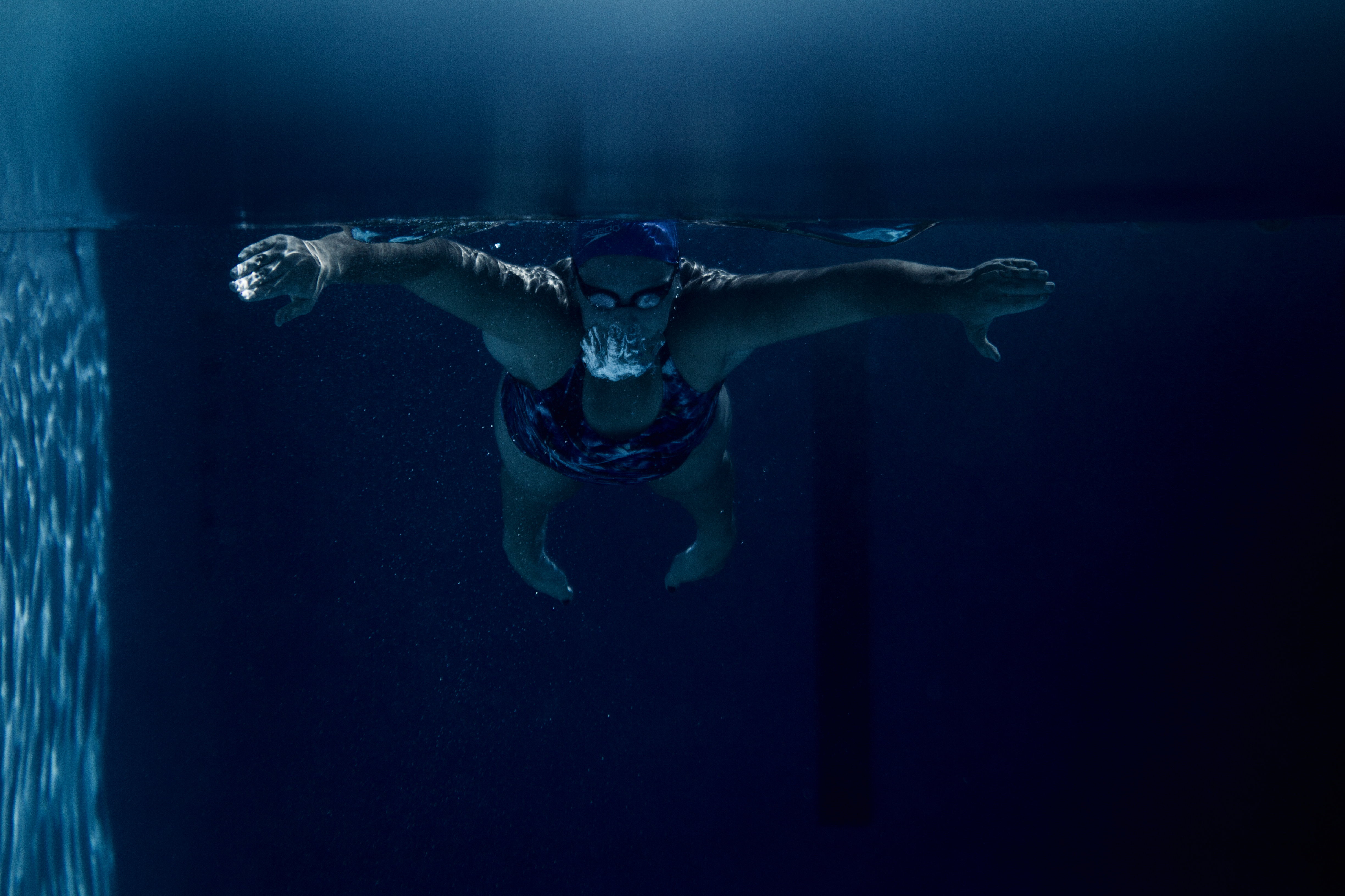 Underwater view of a swimmer wearing a blue cap and goggles, arms outstretched, creating a dynamic contrast in the blue pool.
