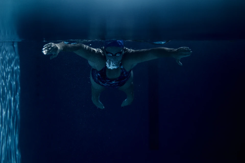 Underwater view of a swimmer wearing a blue cap and goggles, arms outstretched, creating a dynamic contrast in the blue pool.