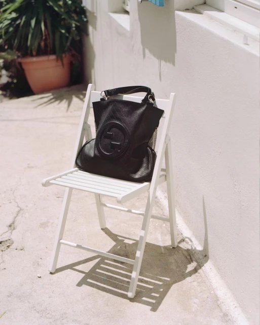 Black leather Gucci handbag displayed on a white folding chair outdoors against a white building, showcasing fashion and design.