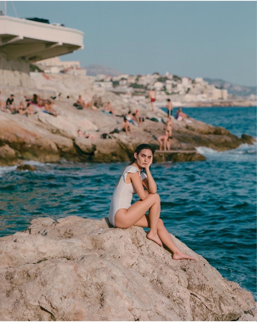 Woman in a white swimsuit poses on a rock formation by the sea on a sunny day with other people relaxing along the coast in background.