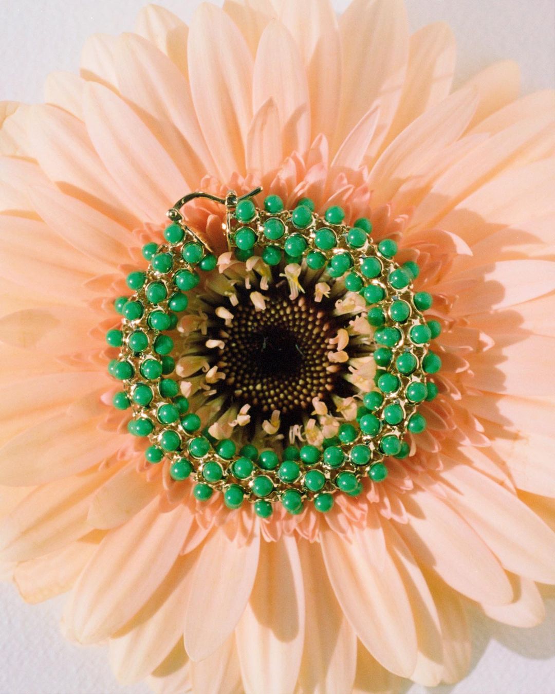 Close-up of a vibrant green and gold beaded earring resting in the center of a soft peach-colored gerbera daisy, highlighting its texture.