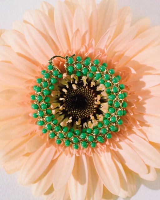 Close-up of a vibrant green and gold beaded earring resting in the center of a soft peach-colored gerbera daisy, highlighting its texture.