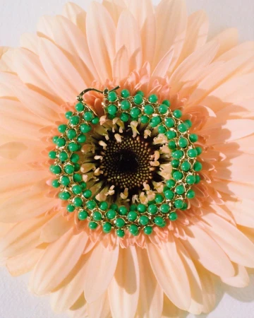 Close-up of a vibrant green and gold beaded earring resting in the center of a soft peach-colored gerbera daisy, highlighting its texture.