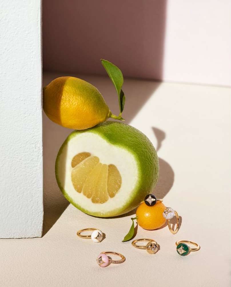 Citrus fruits and gold rings are displayed against a white surface with shadows, evoking luxury and natural beauty in the still life.