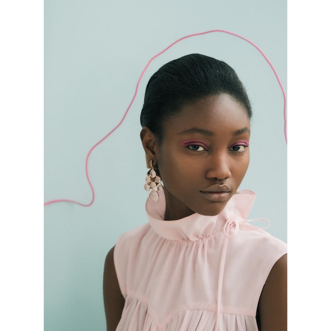 Close-up shot of a dark-skinned woman with pink eye shadow and earrings, wearing a pink ruffled top against a light blue background.