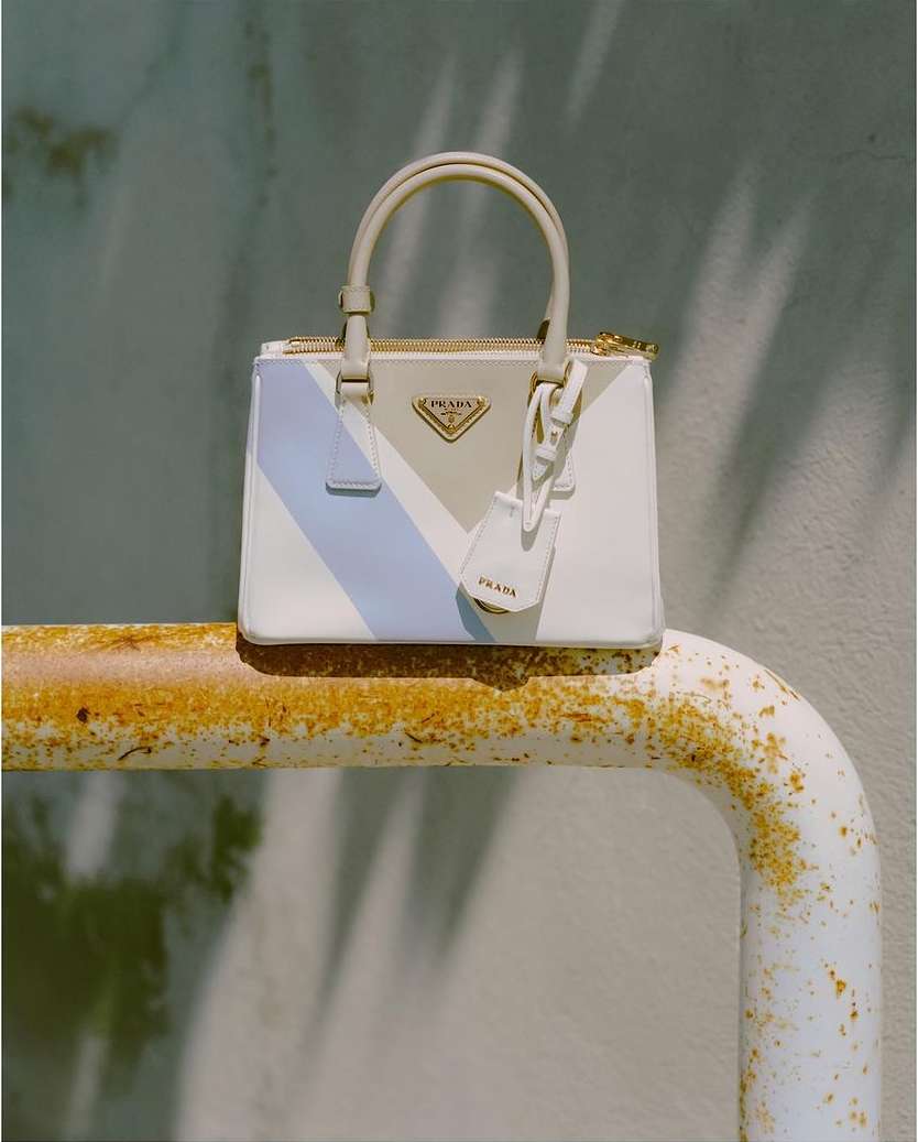 Prada handbag featuring light blue and beige colour-blocking on a weathered metal surface and a concrete wall backdrop with soft shadows.