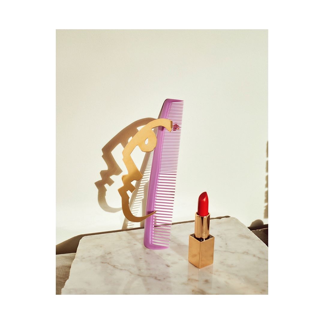 Still life shot featuring a red lipstick tube, light purple comb, and gold abstract shape on a marble surface with soft lighting.