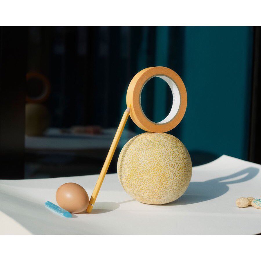 Still life photo of a cantaloupe with an orange tape roll, pencil, egg, and blue wax crayon on a white surface against a dark teal background.