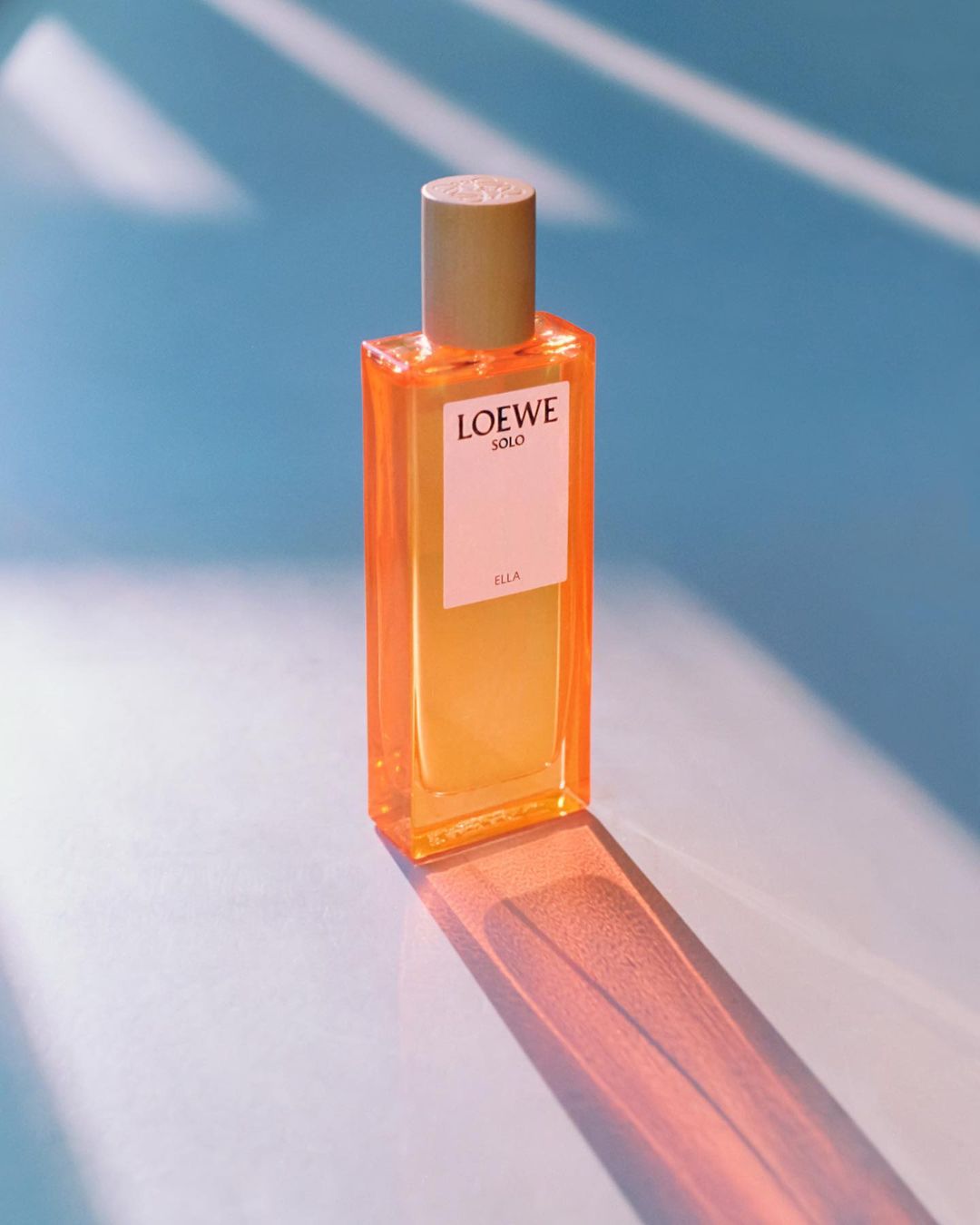 Loewe Solo Ella perfume bottle with golden cap on pastel blue background with geometric shadows, showcasing beauty and elegance.