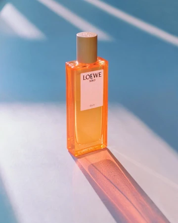 Loewe Solo Ella perfume bottle with golden cap on pastel blue background with geometric shadows, showcasing beauty and elegance.