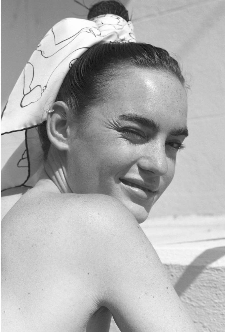 Portrait of a smiling woman with freckles wearing a patterned headscarf, winking, and looking over her shoulder in black and white.