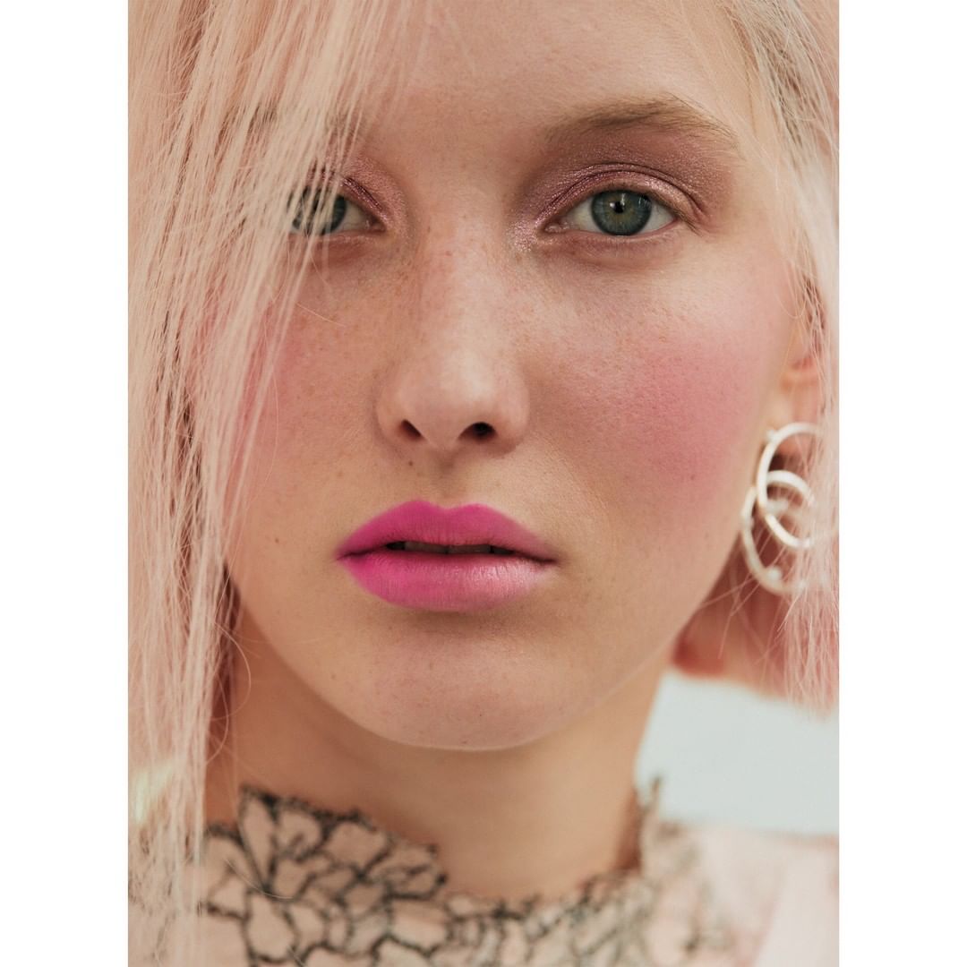 Close-up portrait of a young adult woman with pink hair, silver earrings, pink blush, and bright pink lipstick against a white background.
