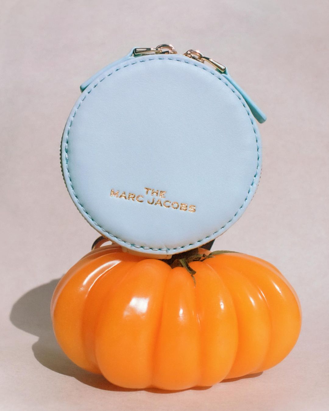 Light blue Marc Jacobs coin purse rests on a bright orange pumpkin in a studio shot with neutral background and soft lighting.