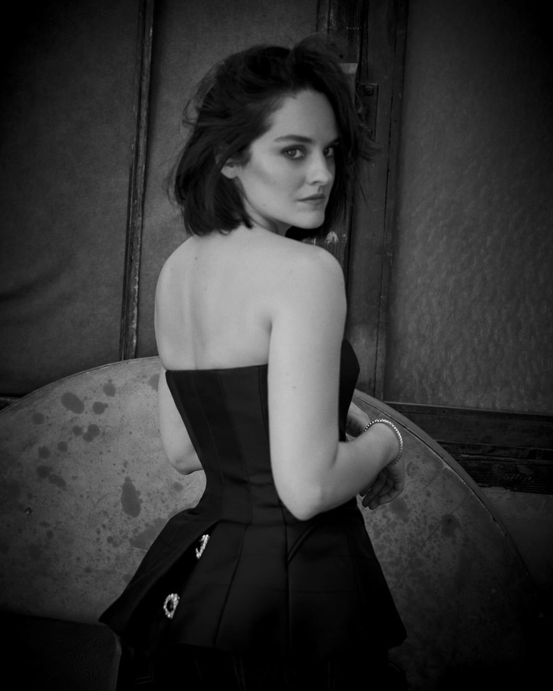 Monochrome portrait of a woman in a strapless black dress looking over her shoulder with loose dark hair against a weathered metal backdrop.