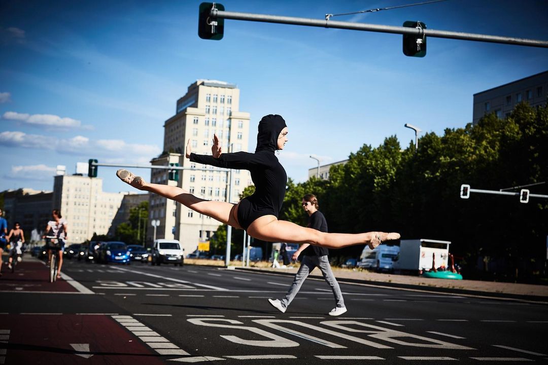 Young adult in athletic wear leaps into a split on a Berlin street during the day with traffic lights, buildings, and pedestrians in background.