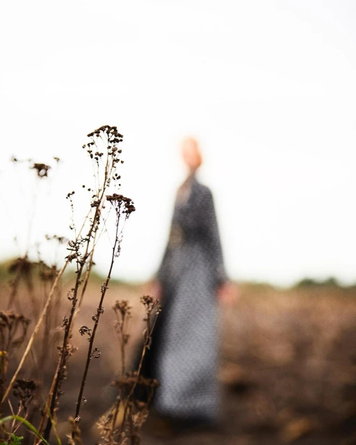 A person in a patterned dress stands in a field of brown grass with dry seed heads in the foreground, all set against a cloudy sky.