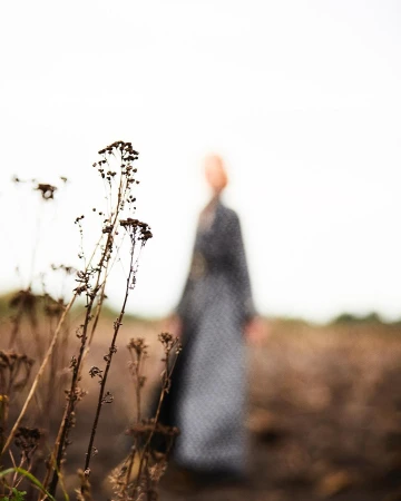 A person in a patterned dress stands in a field of brown grass with dry seed heads in the foreground, all set against a cloudy sky.